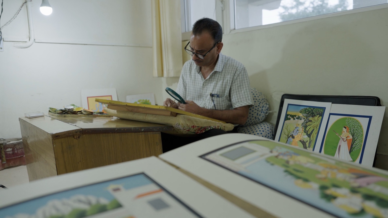 A master painter working with a magnifier at a studio desk surrounded by framed paintings
