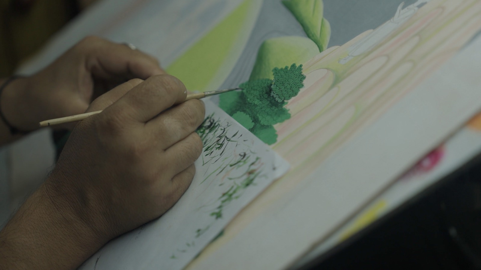Hands steadying a brush to paint foliage in fine green strokes